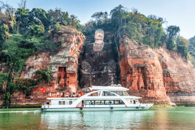 A white sightseeing cruise boat carrying passengers along the river in front of the colossal Leshan Giant Buddha carved into the red cliff face in Sichuan, China.