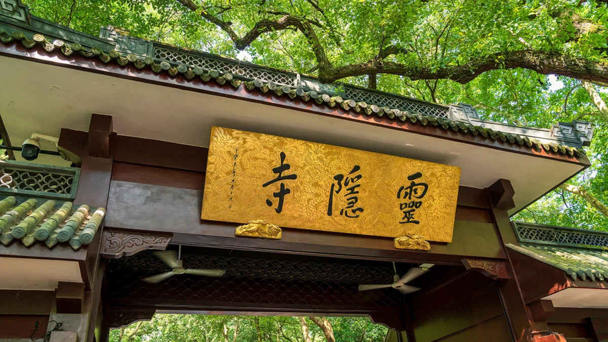 Golden sign board with black Chinese calligraphy at the entrance of Lingyin Temple in Hangzhou, surrounded by dense green trees.