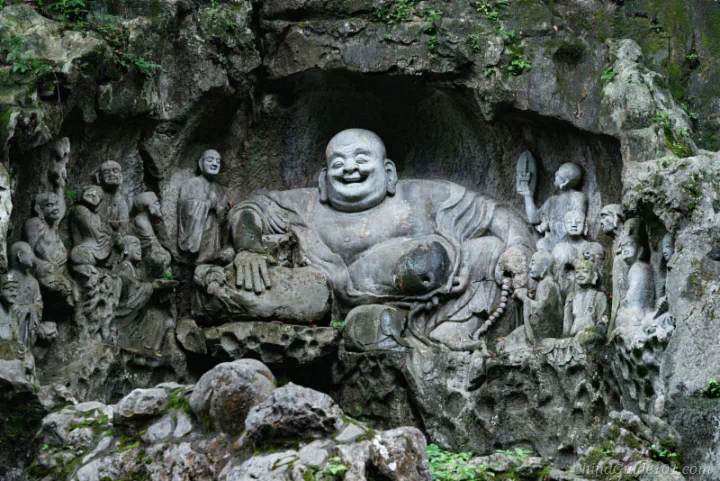 An ancient, large stone carving of the seated laughing Maitreya Buddha with a huge belly, surrounded by smaller attendant figures, set within the rocky cliffside at Feilai Peak, Hangzhou.