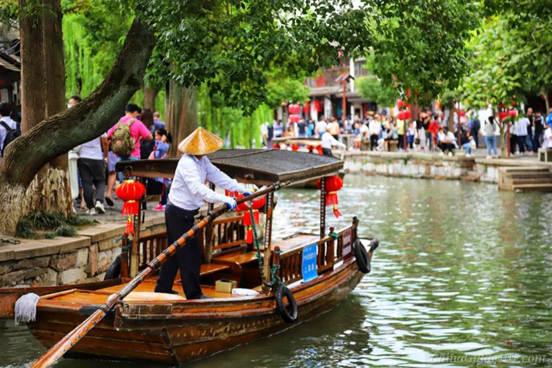 A boatman in a straw hat rows a covered wooden tourist boat decorated with red lanterns on the canal of Zhujiajiao Water Town, Shanghai, with crowds of visitors along the riverside