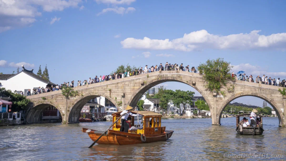Fangsheng Bridge, a five-arch ancient stone bridge in Zhujiajiao Water Town, Shanghai. Tourist wooden rowing boats sail on the river, crowds of visitors stand on the bridge under blue sky with white clouds