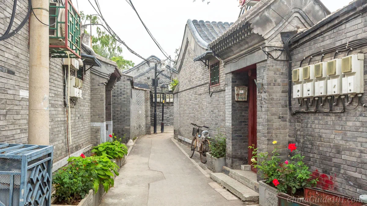 Traditional Beijing hutong alley with grey-brick courtyard walls, red doorways, flower pots and a bicycle leaning against the wall
