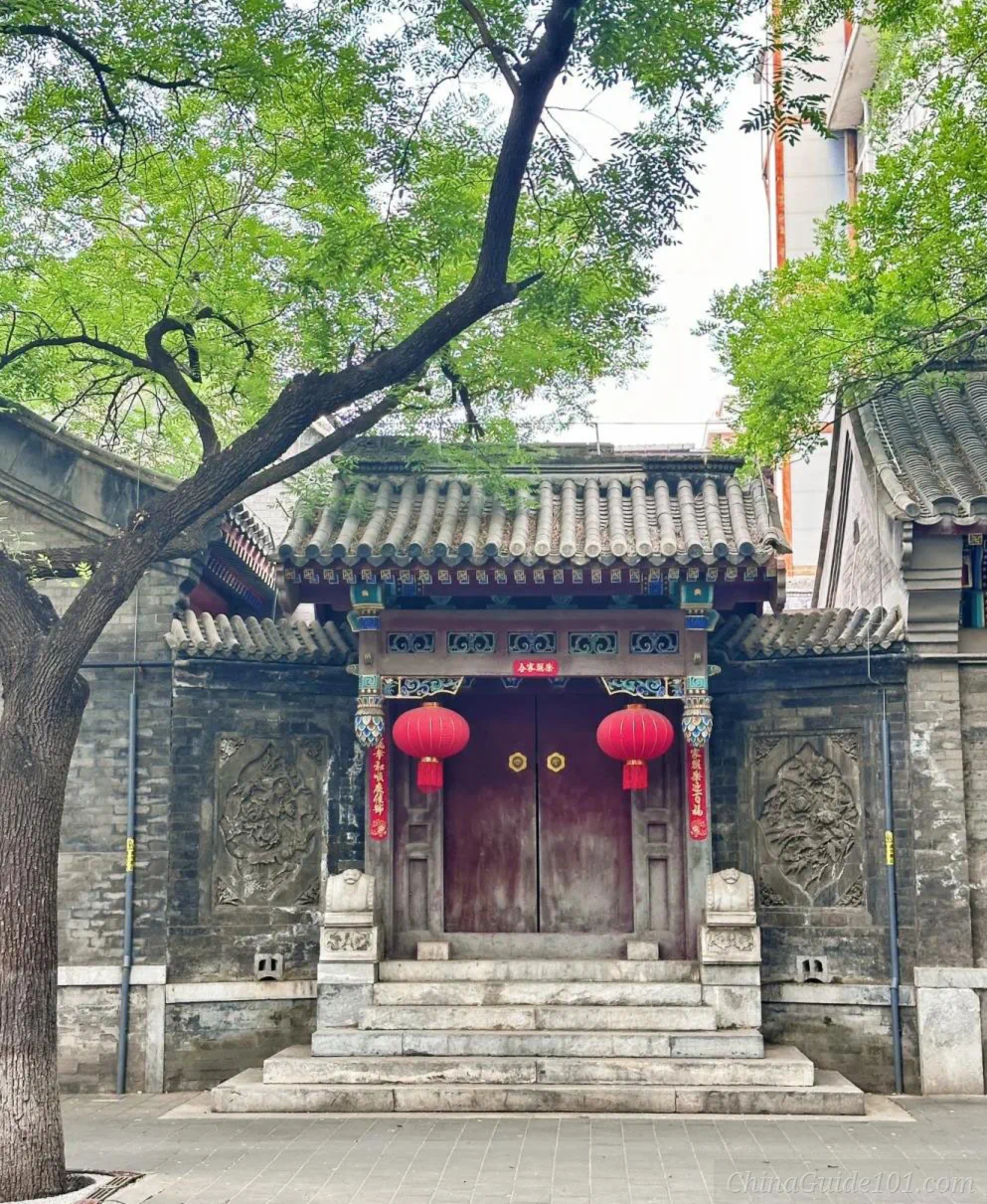 Traditional Beijing siheyuan courtyard gate with stone door stools, grey brick carvings, tiled eaves and red lanterns under a locust tree