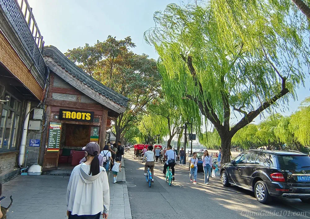 Shichahai scenic area in Beijing, bustling street beside the lake, old-style shops, willow trees, cyclists and walking visitors